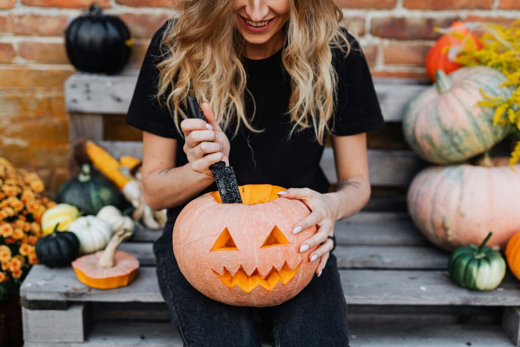 A cheerful woman carves a jack-o'-lantern amidst fall pumpkins on a wooden bench, celebrating Halloween.