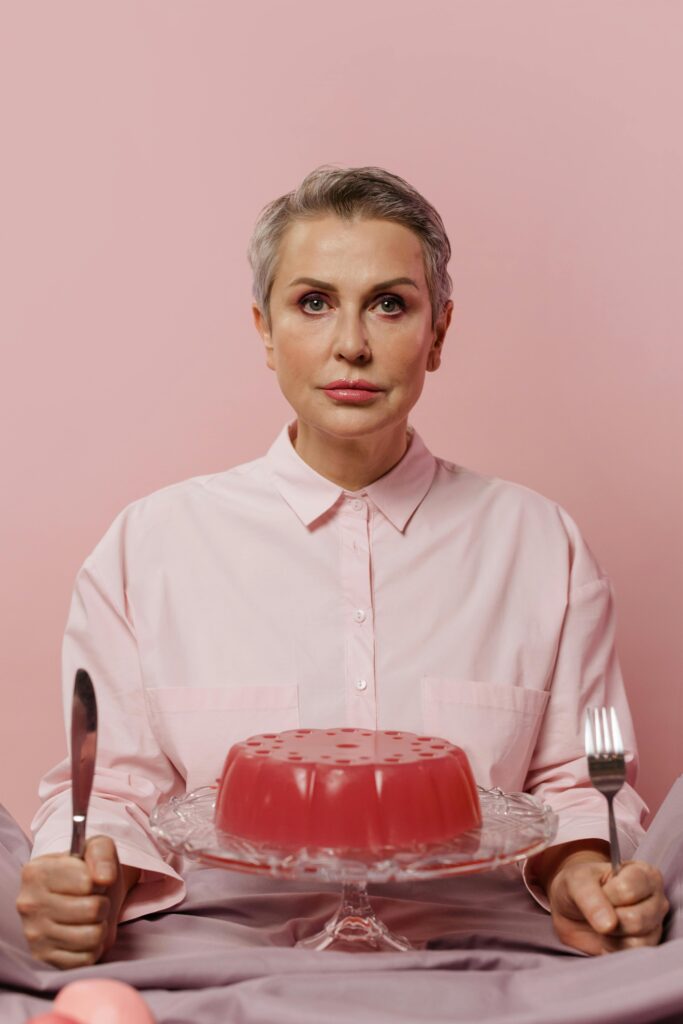 Portrait of a woman with short gray hair holding a knife and fork, seated with a pink jelly dessert.