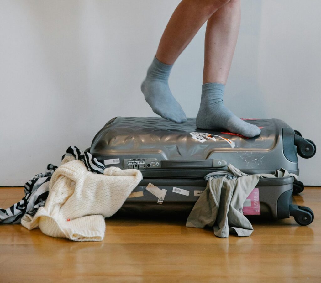 A woman attempts to close an overloaded suitcase by standing on it, surrounded by clothes.